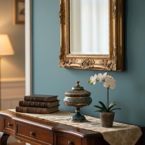 Könnte beinhalten: Eine Holzkonsole mit drei Schubladen, verziert mit einem dekorativen Läufer, Büchern und einem Deckelgefäß. Eine Topf-Orchidee und ein goldgerahmter Spiegel hängen an der türkisblauen Wand und schaffen ein elegantes Interieur.