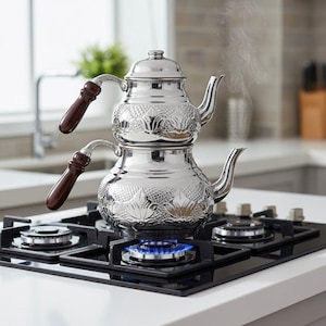 May include: A two-tiered, stainless steel teapot with ornate leaf patterns and dark brown wooden handles. The teapot is steaming on a black gas stovetop with a blue flame, suggesting it is in use. The kitchen setting includes a white countertop and a blurred background.