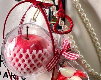 May include: A clear spherical ornament containing a red apple wrapped in a white protective foam net, accented with a red gingham ribbon. Beside it, a small clear bag holds red and white apple-shaped charms, finished with a pearl bead chain.