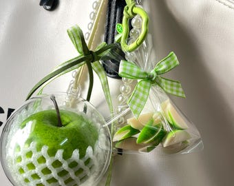 May include: A bag charm featuring a clear spherical ornament containing a green apple wrapped in white foam netting, alongside a small clear plastic bag filled with green apple slices, both accented with green gingham ribbons.