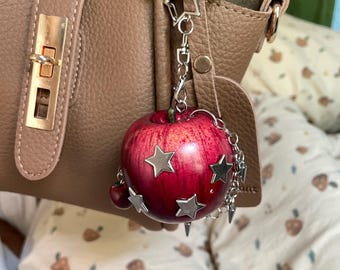 May include: A red apple-shaped charm hanging from a brown leather handbag. The apple features metallic silver star-shaped embellishments and is attached to a silver-toned chain with a star-shaped clasp and small dangling metal spikes.