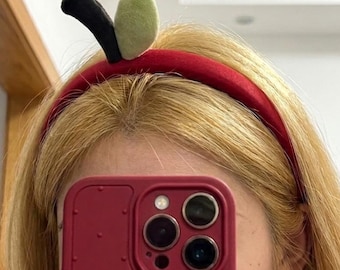 Può includere: Un cerchietto in velluto rosso con un picciolo di mela in peluche e una singola foglia verde chiaro sulla parte superiore. Il cerchietto è indossato sopra capelli biondi, posizionato sopra una cover per smartphone rosso scuro con superficie puntinata.