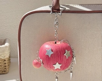 May include: A bag charm featuring a pink apple decorated with silver star-shaped accents and small metal spikes. The charm is attached to a silver chain with a star-shaped clasp, hanging from the brown handle of a beige textured handbag.