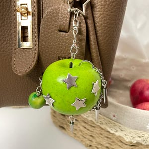 May include: A bright green apple charm attached to a silver-toned metal keychain. The apple features several silver star-shaped embellishments, a small dangling green apple charm, and multiple silver metal spikes hanging from a delicate chain.