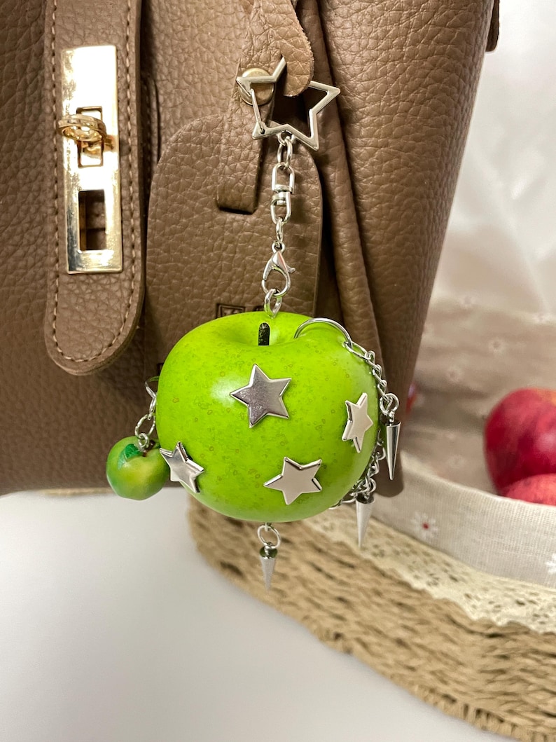 May include: A bright green apple charm attached to a silver-toned metal keychain. The apple features several silver star-shaped embellishments, a small dangling green apple charm, and multiple silver metal spikes hanging from a delicate chain.