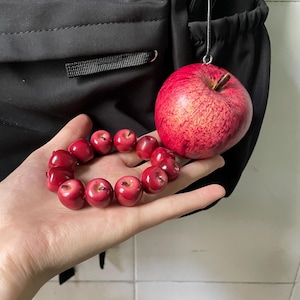 Puede incluir: Una pulsera compuesta por pequeñas cuentas rojas brillantes con forma de manzana y un dije de manzana roja realista con tallo marrón, unido a un cordón negro. El dije de manzana presenta una piel texturizada con variaciones de color.