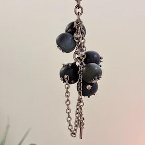 May include: A silver-toned metal chain necklace featuring a cluster of dark blue, spherical berry-like charms. Each sphere is accented with small, sharp metallic spikes and embedded clear crystals, suspended from a delicate link chain structure.