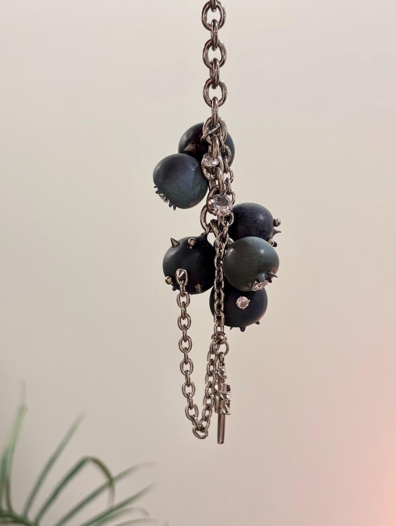 May include: A silver-toned metal chain necklace featuring a cluster of dark blue, spherical berry-like charms. Each sphere is accented with small, sharp metallic spikes and embedded clear crystals, suspended from a delicate link chain structure.