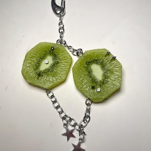 May include: A keychain featuring two realistic kiwi fruit slices attached to a silver-toned metal chain. Each kiwi slice is decorated with small metal spikes and clear rhinestones. The chain includes two dangling silver-toned star charms and a lobster clasp.