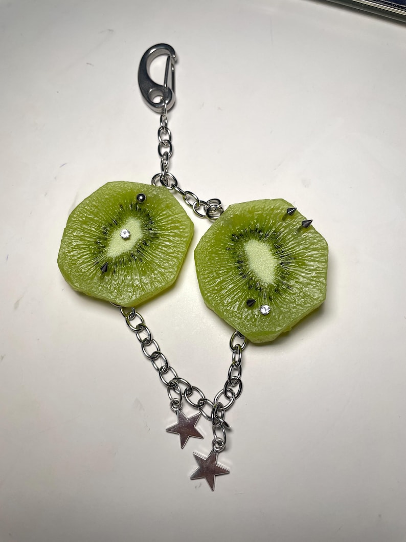 May include: A keychain featuring two realistic kiwi fruit slices attached to a silver-toned metal chain. Each kiwi slice is decorated with small metal spikes and clear rhinestones. The chain includes two dangling silver-toned star charms and a lobster clasp.