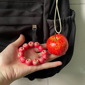 May include: A bracelet composed of small, round, red apple-shaped beads with tiny green leaves, alongside a larger, realistic red apple charm featuring yellow streaks and a brown stem, attached to a light-colored cord loop for hanging.