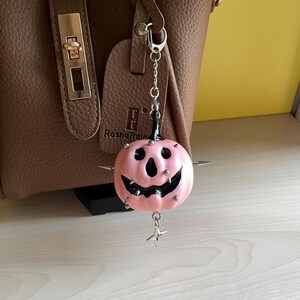 May include: A pink pumpkin-shaped charm with a black carved face, silver metal spikes, and a dangling star-shaped pendant. The charm is attached to a silver chain with a lobster clasp, hanging in front of a brown textured handbag with gold hardware.