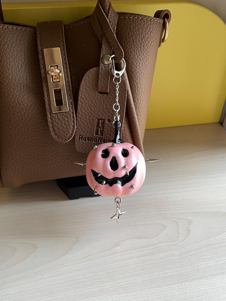 May include: A pink pumpkin-shaped charm with a black carved face, silver metal spikes, and a dangling star-shaped pendant. The charm is attached to a silver chain with a lobster clasp, hanging in front of a brown textured handbag with gold hardware.