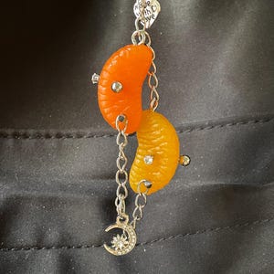May include: A silver-toned metal chain keychain featuring two orange citrus segments, one bright orange and one yellow-orange, each accented with small silver studs and clear rhinestones. A heart-shaped charm with the text Made with Love and a crescent moon charm with a sun center are attached to the chain.