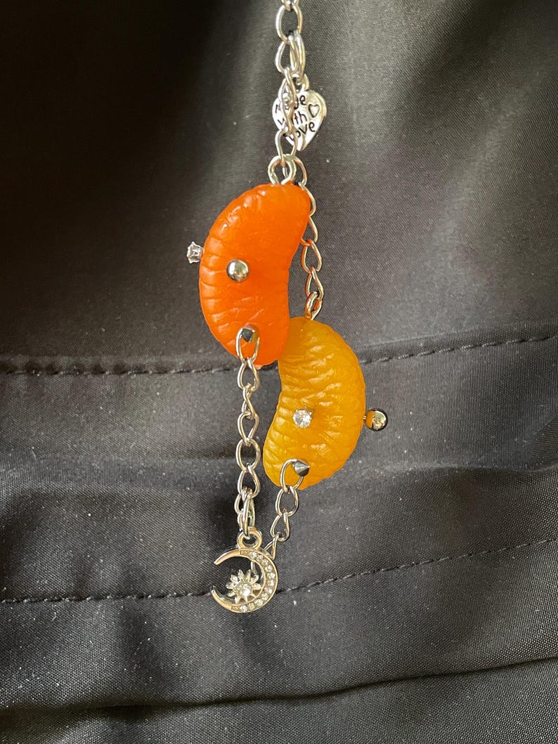 May include: A silver-toned metal chain keychain featuring two orange citrus segments, one bright orange and one yellow-orange, each accented with small silver studs and clear rhinestones. A heart-shaped charm with the text Made with Love and a crescent moon charm with a sun center are attached to the chain.