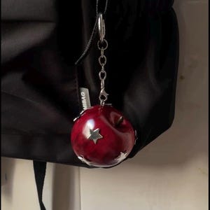 May include: A round, glossy, deep red apple-shaped charm featuring metallic silver star accents and a silver-toned metal chain attachment. The charm is suspended from a black fabric bag, with a small white tag visible reading MIBAG.