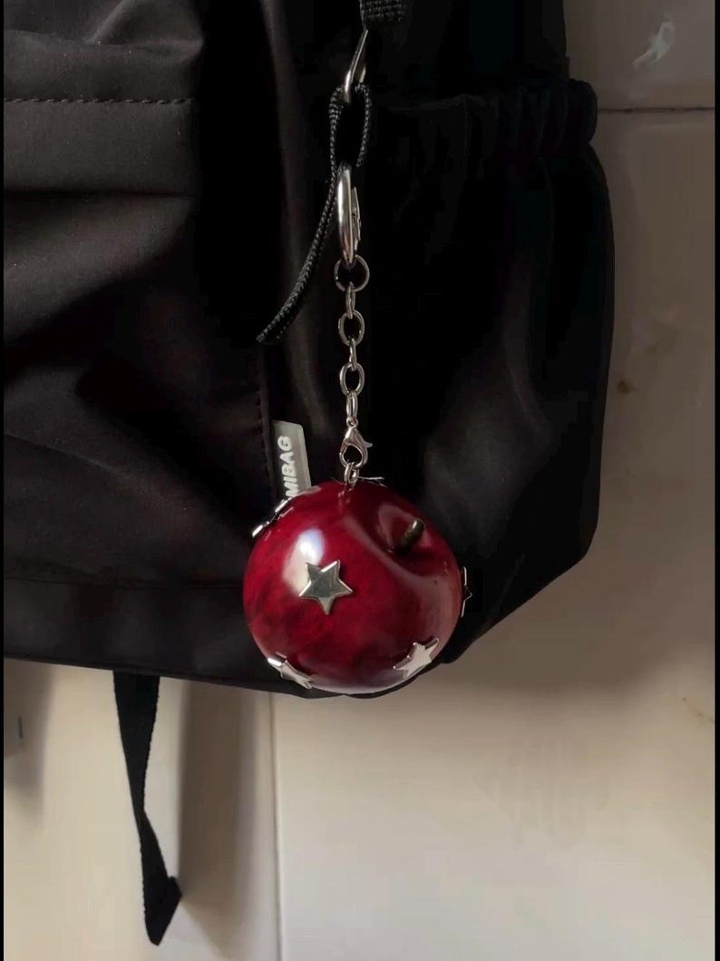 May include: A round, glossy, deep red apple-shaped charm featuring metallic silver star accents and a silver-toned metal chain attachment. The charm is suspended from a black fabric bag, with a small white tag visible reading MIBAG.