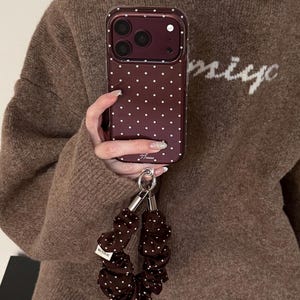 May include: A dark maroon smartphone case featuring a white polka dot pattern and a matching dark brown fabric scrunchie wrist strap with white polka dots. The case has a camera cutout and a small silver logo at the bottom, with a silver metal ring attachment.