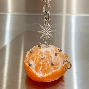 May include: A keychain featuring a peeled orange segment adorned with small silver-toned metal spikes and studs. A silver-toned star-shaped charm with embedded clear crystals hangs from the top chain, which includes a lobster claw clasp.