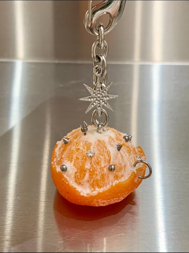 May include: A keychain featuring a peeled orange segment adorned with small silver-toned metal spikes and studs. A silver-toned star-shaped charm with embedded clear crystals hangs from the top chain, which includes a lobster claw clasp.