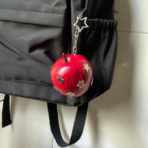 May include: A red apple-shaped charm with a brown stem and silver star-shaped accents attached to a silver star-shaped clasp. The charm hangs from the side of a black backpack, featuring a smooth, rounded surface with multiple metallic star embellishments.