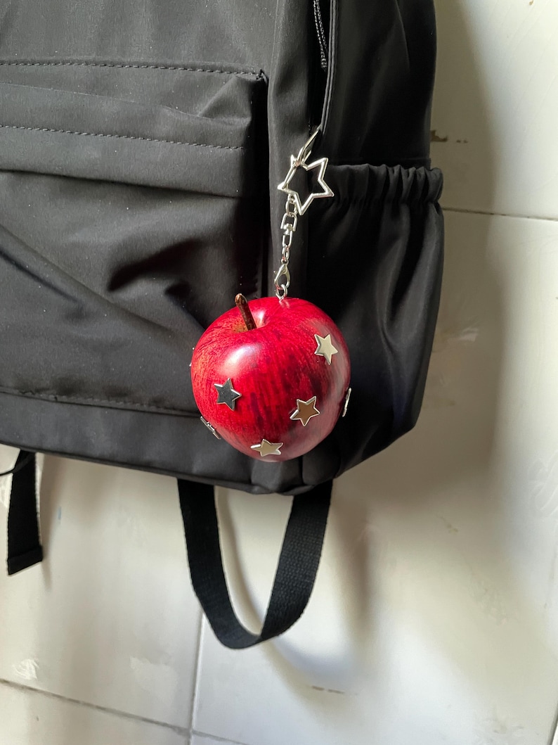 May include: A red apple-shaped charm with a brown stem and silver star-shaped accents attached to a silver star-shaped clasp. The charm hangs from the side of a black backpack, featuring a smooth, rounded surface with multiple metallic star embellishments.