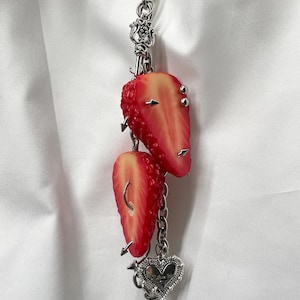 May include: A silver-toned metal chain charm featuring two realistic strawberry slices adorned with small silver spikes and rings. The accessory includes a decorative silver heart-shaped pendant at the bottom, creating a detailed piece of jewelry.