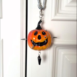 May include: A pumpkin-shaped keychain featuring a gradient orange and yellow finish with a black stem and carved face. The pumpkin is adorned with silver metal spikes and studs, and a small black coffin-shaped charm hangs from the bottom.