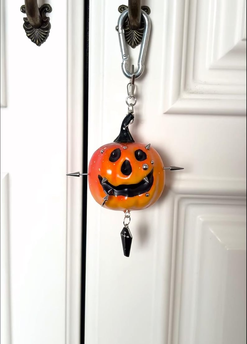 May include: A pumpkin-shaped keychain featuring a gradient orange and yellow finish with a black stem and carved face. The pumpkin is adorned with silver metal spikes and studs, and a small black coffin-shaped charm hangs from the bottom.