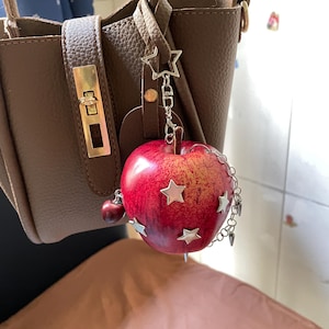 May include: A red apple-shaped charm hanging from a brown leather handbag. The apple features silver-toned star-shaped embellishments, a small dangling cherry charm, and a silver chain with small spikes attached to the side of the fruit.