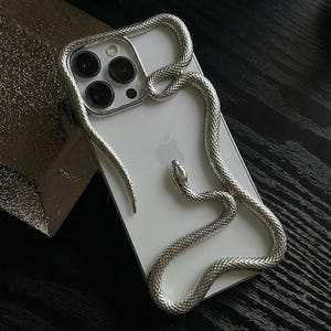 以下が含まれることがあります： 背面に立体的なメタリックシルバーの蛇が巻き付いた白いスマートフォンケース。蛇の質感のある胴体は、上部のカメラモジュールからデバイスの中央に向かって伸びており、詳細な鱗の模様が施されています。