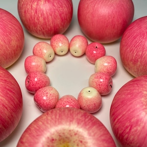 Puede incluir: Una composición de manzanas rojas y objetos más pequeños con forma de manzana de color rojo rosado. Las manzanas más grandes tienen un tallo oscuro y una superficie texturizada. Los objetos más pequeños están dispuestos en círculo, rodeados por las manzanas más grandes.