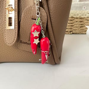 May include: A silver-toned metal keychain featuring two bright red chili pepper charms with green stems. The peppers are decorated with small silver star-shaped accents and pointed metal studs, all suspended from a star-shaped metal ring clasp.