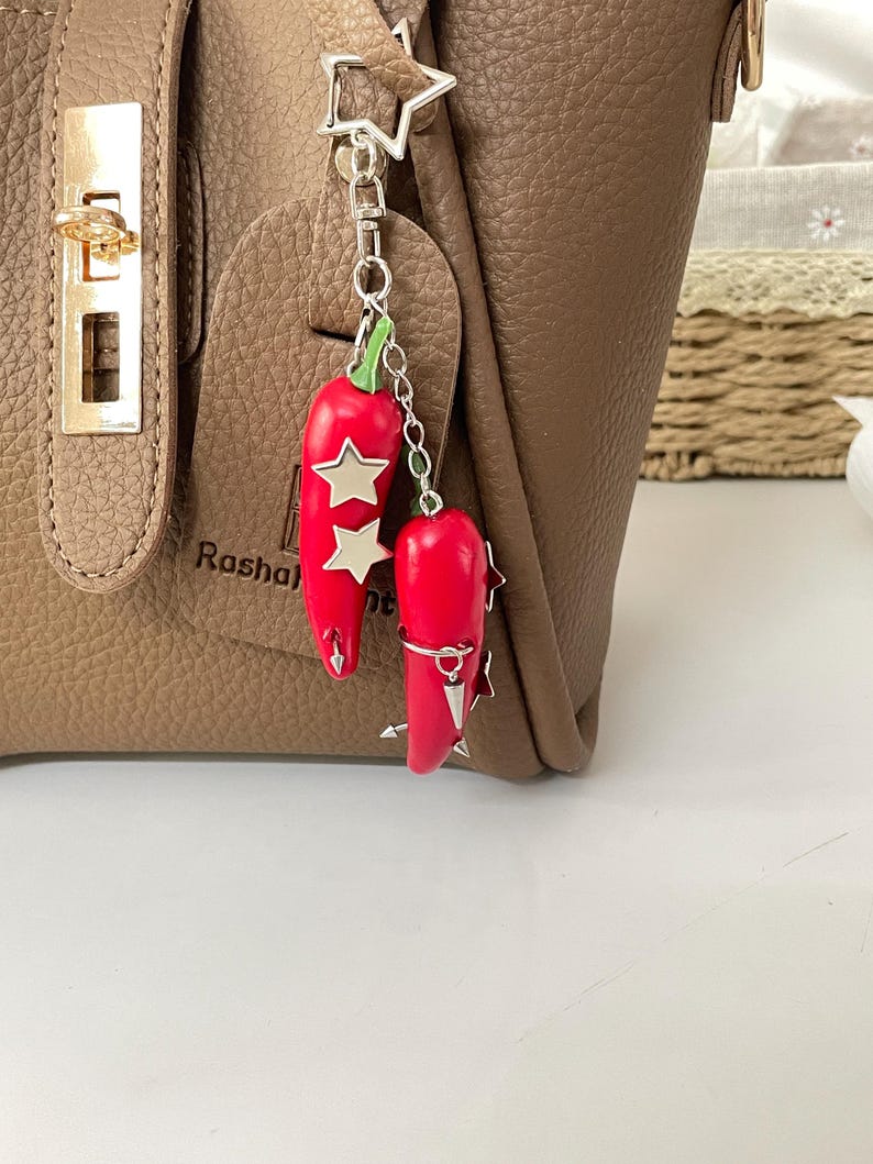 May include: A silver-toned metal keychain featuring two bright red chili pepper charms with green stems. The peppers are decorated with small silver star-shaped accents and pointed metal studs, all suspended from a star-shaped metal ring clasp.