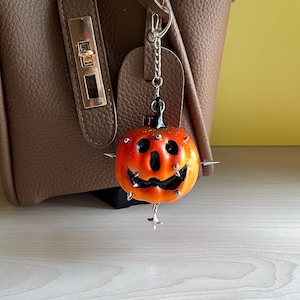 May include: A pumpkin-shaped charm featuring a gradient orange and yellow finish with a black carved face. The charm is adorned with small silver-toned metal spikes and studs, and it hangs from a silver-toned metal chain with a lobster claw clasp.