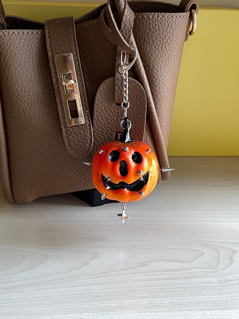 May include: A pumpkin-shaped charm featuring a gradient orange and yellow finish with a black carved face. The charm is adorned with small silver-toned metal spikes and studs, and it hangs from a silver-toned metal chain with a lobster claw clasp.