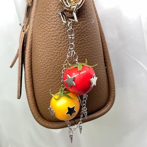 May include: A bag charm featuring a red tomato and a yellow tomato, each adorned with silver-toned star-shaped accents and green stems. The charms are attached to a silver-toned chain with a star-shaped clasp and small metal spike embellishments.