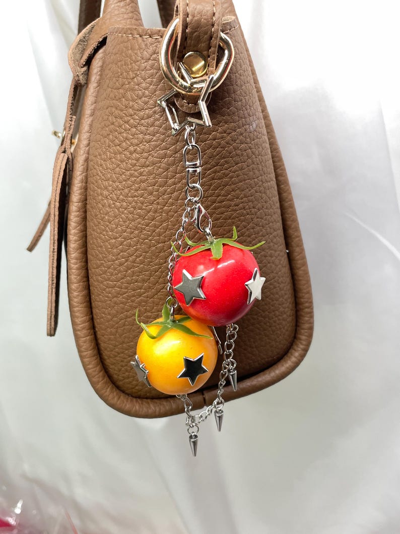 May include: A bag charm featuring a red tomato and a yellow tomato, each adorned with silver-toned star-shaped accents and green stems. The charms are attached to a silver-toned chain with a star-shaped clasp and small metal spike embellishments.