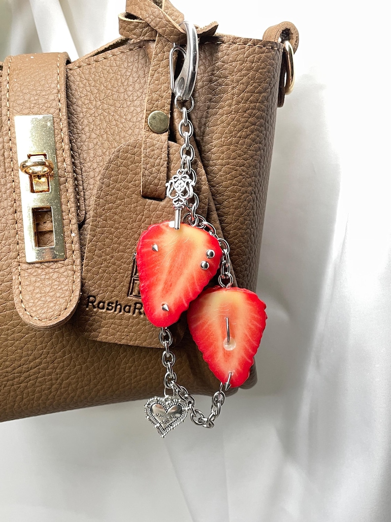 May include: A silver-toned metal keychain featuring two realistic strawberry slices and a small heart charm attached to a chain. The keychain is clipped onto a textured brown leather handbag that has a gold-toned metal clasp and the text RashaR.