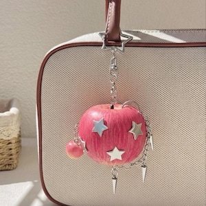 May include: A pink apple-shaped charm hanging from a brown handbag strap. The charm features silver star-shaped accents, a small dangling pink sphere, and silver chain links with pointed metal spikes attached to the side of the apple.