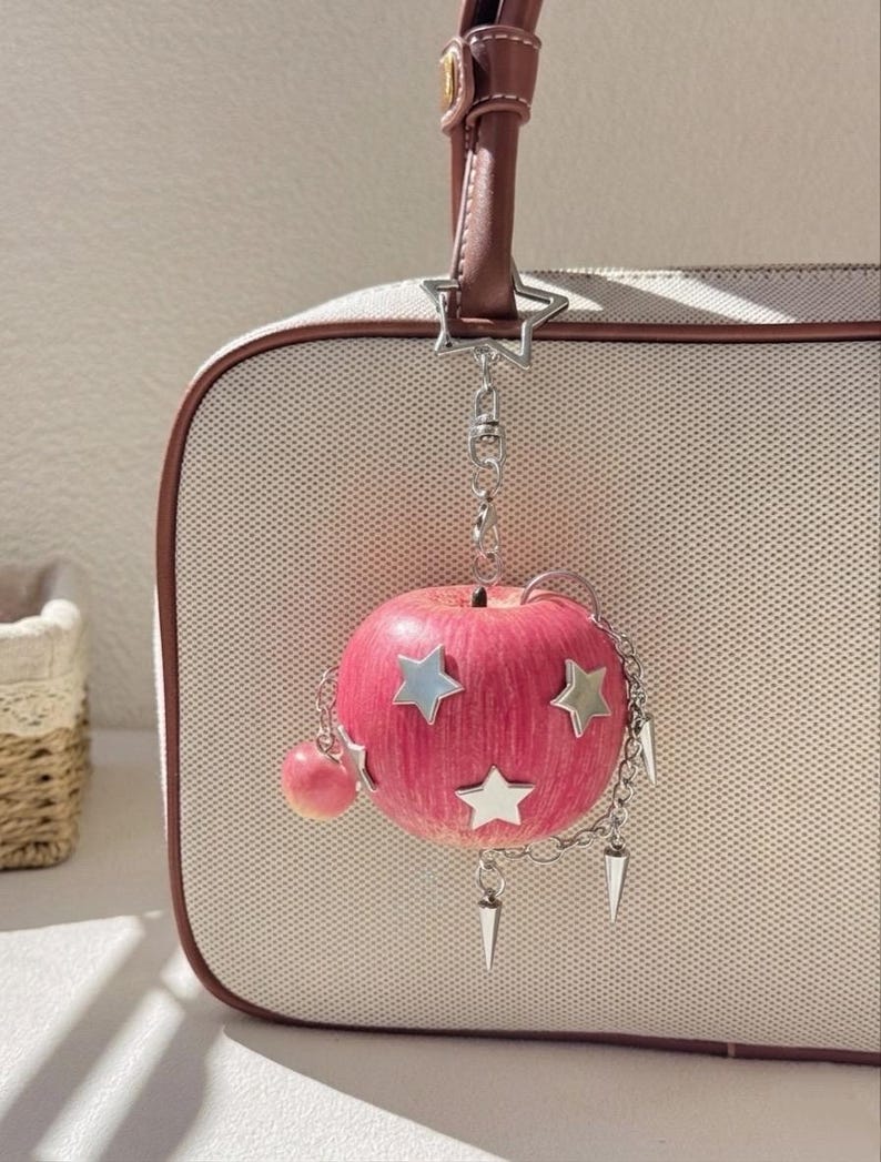 May include: A pink apple-shaped charm hanging from a brown handbag strap. The charm features silver star-shaped accents, a small dangling pink sphere, and silver chain links with pointed metal spikes attached to the side of the apple.