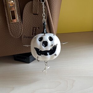 May include: A white pumpkin-shaped charm with a black painted face, featuring silver-toned metal spikes and studs embedded into the surface. The charm hangs from a silver-toned metal chain with a small star-shaped pendant attached at the bottom.
