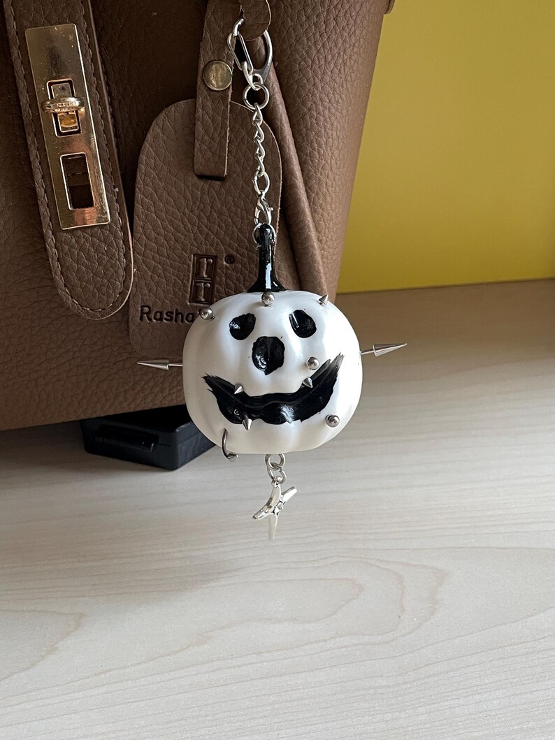 May include: A white pumpkin-shaped charm with a black painted face, featuring silver-toned metal spikes and studs embedded into the surface. The charm hangs from a silver-toned metal chain with a small star-shaped pendant attached at the bottom.