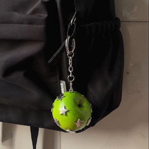 May include: A bright green apple-shaped charm attached to a silver-toned metal chain and ring. The apple features several small, metallic silver star-shaped embellishments fixed to its surface. The charm is suspended from a black fabric backpack.