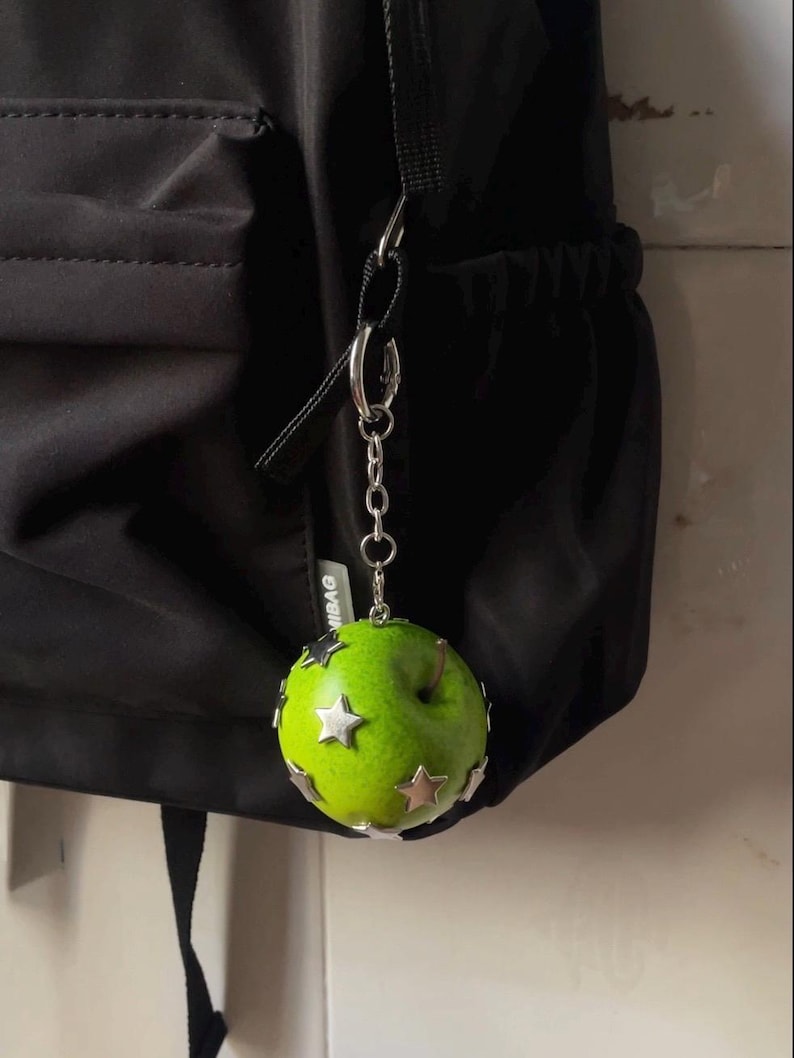 May include: A bright green apple-shaped charm attached to a silver-toned metal chain and ring. The apple features several small, metallic silver star-shaped embellishments fixed to its surface. The charm is suspended from a black fabric backpack.