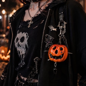May include: A black zip-up hoodie featuring a distressed knit top with a white skull graphic. The hoodie is adorned with a silver bat pin, a star-shaped safety pin, and a hanging orange pumpkin charm accented with small silver studs and a black crystal.