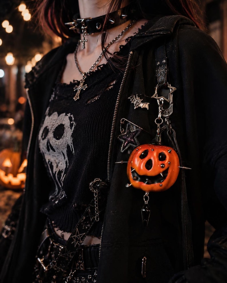 May include: A black zip-up hoodie featuring a distressed knit top with a white skull graphic. The hoodie is adorned with a silver bat pin, a star-shaped safety pin, and a hanging orange pumpkin charm accented with small silver studs and a black crystal.