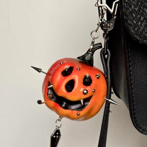 May include: A pumpkin-shaped charm featuring a gradient orange and red finish with a carved, smiling face. The pumpkin is decorated with multiple silver-toned metal spikes and studs, and it hangs from a silver-colored star-shaped metal clasp.