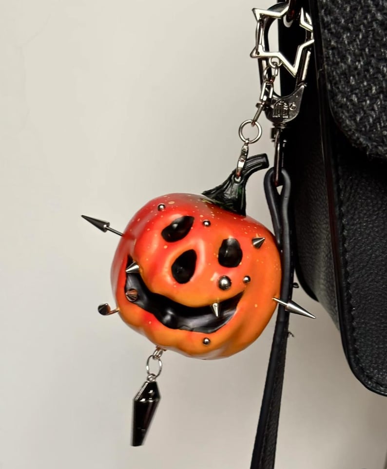 May include: A pumpkin-shaped charm featuring a gradient orange and red finish with a carved, smiling face. The pumpkin is decorated with multiple silver-toned metal spikes and studs, and it hangs from a silver-colored star-shaped metal clasp.