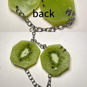 May include: A necklace featuring two green kiwi fruit slice pendants connected by a silver-toned metal chain. The pendants are embellished with small metal spikes and clear rhinestones. The word back is printed between the two pendants.